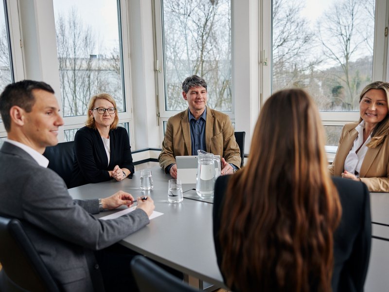 Im Vordergrund sitzen ein Geschäftsführer und eine Mitarbeiterin mit dem Rücken zur Kamera. Gegenüberliegend sieht man den 2. Geschäftsführer und links und rechts von ihm 2 weitere Mitarbeiterinnen.